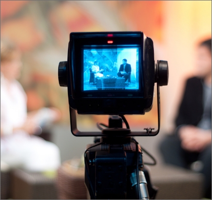 A video camera recording a unfocused interview between two people in the front. The view of the camera is from the back.